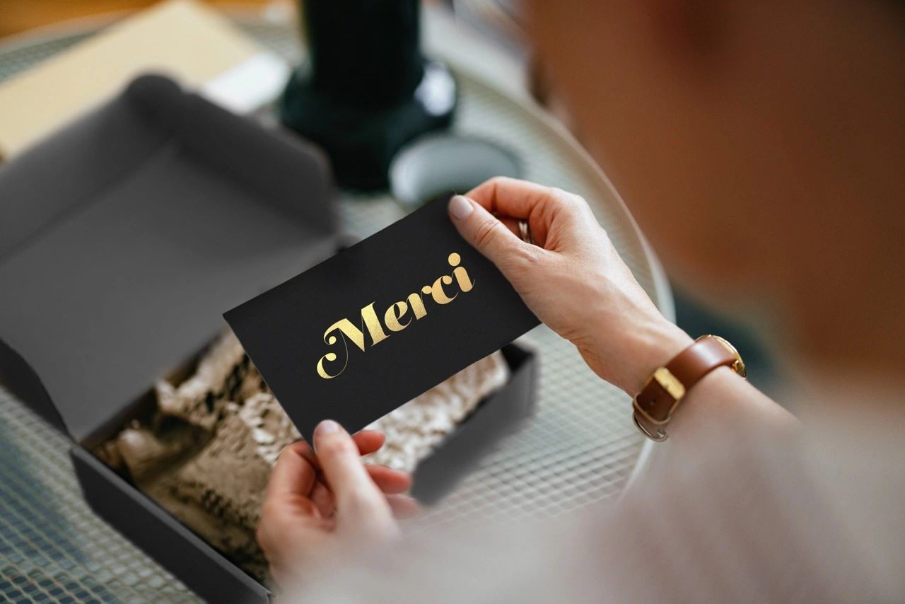 une femme sort d'une boîte cadeau une carte noire avec une inscription dorée "merci".